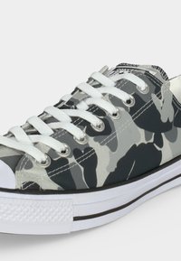 Χαμηλό sneaker με γκρι και μαύρο καμουφλάζ σχεδιασμό, λευκά κορδόνια, λευκή ελαστική μύτη και λευκή σόλα με μαύρη ρίγα.