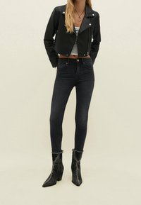 Chaqueta corta de cuero sintético negro con herrajes plateados, camiseta sin mangas gris, vaqueros ajustados oscuros y botines negros texturizados con tacones de bloque.