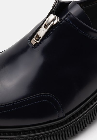 Navy blue leather shoe with a glossy finish, featuring a silver zipper along the front and textured black sole for grip.