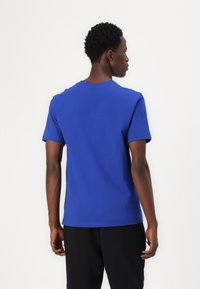 T-shirt de algodão azul, com gola redonda e mangas curtas. Textura suave com um design simples. Usada com calças pretas, vista de costas.