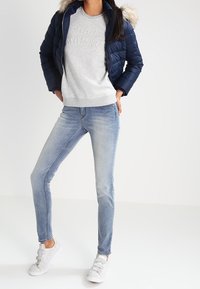 Granatowa kurtka puchowa z kapturem wykończonym futrem, szary sweatshirt z wypukłym napisem, jasnoniebieskie rurki i białe trampki na rzepy.