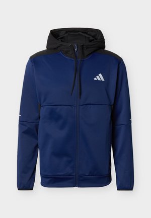 Chaqueta azul marino con cremallera, capucha negra, paneles negros en los hombros y logotipo blanco en el pecho izquierdo, presentada sobre un fondo liso.