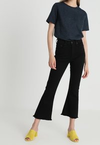 Sorte flarejeans med rå kant; kombineret med en mørk, plettet kortærmet top. Gule mule-sko med åbne tæer fuldender outfit.
