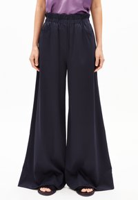 Weite, marineblaue Hosen mit elastischem Bund, seitlichen Taschen und glatter Textur, kombiniert mit schwarzen, offenen Schuhen.