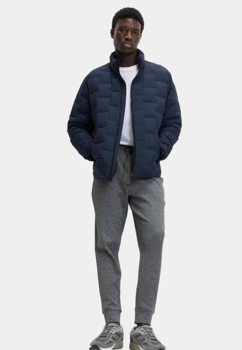 Chaqueta acolchada azul marino con un cuello alto y cremallera frontal, usada sobre una camisa blanca, combinada con pantalones de chándal grises y zapatillas grises.