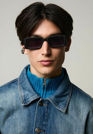 Lunettes de soleil rectangulaires noires avec des verres sombres, portées sur un pull à col roulé bleu à côtes, associées à une veste en denim.