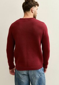 Czerwony sweter z dzianiny o teksturowanym wzorze, okrągłym dekolcie oraz ściągaczami przy mankietach i u dołu. Zestawiony z jasnoniebieskimi dżinsami.