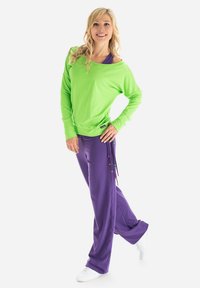 Un haut vert à manches longues avec un décolleté large s'associe à un pantalon violet doté d'accents colorés en cordon de serrage et à des chaussures de sport blanches.