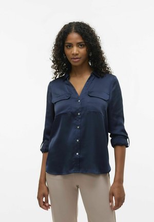Camicia button-up in satin blu navy con due tasche frontali, maniche arrotolate e colletto. Texture liscia con una leggera lucentezza.