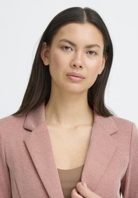 Blazer rosa claro con tela texturizada, solapas con muescas y diseño ajustado. Se lleva sobre una blusa marrón, que presenta un escote abierto.