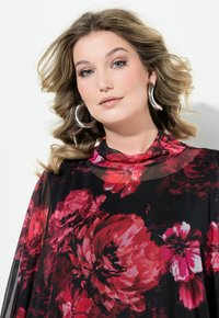 Blusa nera a fiori con grandi fiori rossi, collo alto e maniche trasparenti. Gli accessori includono grandi orecchini a cerchio in argento.