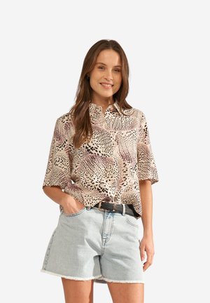 Woman with long brown hair wearing a patterned short-sleeve blouse, light denim shorts, and a black belt, smiling with one hand in pocket.