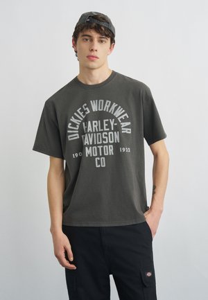 Jeune homme portant un t-shirt gris foncé "Dickies Workwear Harley-Davidson Motor Co" et un pantalon cargo noir, avec une casquette noire portée à l'envers.