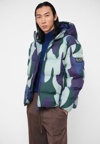 Homem com uma jaqueta acolchoada azul com padrão e capuz, gola alta azul-marinho, e calças castanhas, de pé com as mãos nos bolsos contra um fundo branco.