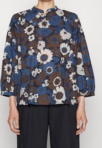 Blusa con motivo floreale caratterizzata da fiori blu, marroni e bianchi, maniche lunghe, colletto a fascia e vestibilità comoda. Tessuto texturizzato.