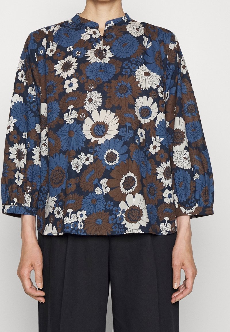 Blusa con motivo floreale caratterizzata da fiori blu, marroni e bianchi, maniche lunghe, colletto a fascia e vestibilità comoda. Tessuto texturizzato.