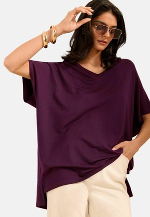 Egy nő arany karkötőket és napszemüveget igazít, laza lila V-nyakú felsőt és világos bézs nadrágot visel, egyszerű háttér előtt.