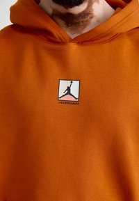 Haut à capuche orange en mélange de coton, avec un petit logo Jumpman noir et rose accompagnant le texte "JORDAN", présentant une texture lisse.