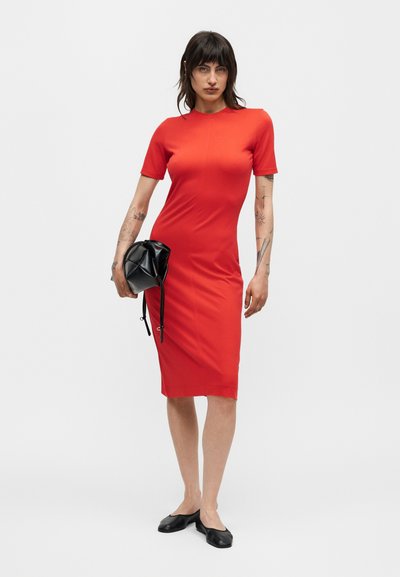 Γυναίκα με σκουρόχρωμα μαλλιά που φοράει εφαρμοστή κόκκινη φορεσιά μέχρι το γόνατο, κρατώντας μια μαύρη τσάντα χειρός στο αριστερό της χέρι, φορώντας μαύρα επίπεδα παπούτσια, στέκεται σε λευκό φόντο.