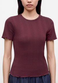 Femme portant un haut bordeaux côtelé ajusté à manches courtes avec des bords en forme de laitue et une jupe foncée sur un fond uni.