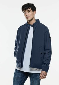 Chaqueta ligera azul marino con cuello alto, cremallera frontal, detalles de botones a presión y bolsillos laterales, combinada con una camiseta blanca.