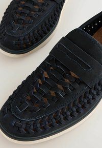 Mocasines tejidas de color azul marino, con parte superior de gamuza, costuras de acento y una suela de goma ligera. Incluyen una correa decorativa en el empeine.
