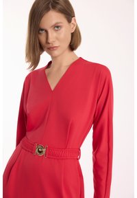 Czerwona sukienka z gładkiego materiału, z długimi rękawami, dekoltem w kształcie litery V i paskiem w talii z okrąłym złotym akcentem.