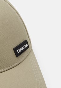 Cappellino da baseball khaki con forma strutturata, dotato di un'etichetta rettangolare nera con la scritta "Calvin Klein" in bianco e dettagli di cucitura visibili.