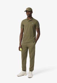 Polo shirt verde oliva con logo, pantaloni jogger abbinati. Sneakers chiare, tiene in mano una pallina da tennis gialla. Il cappellino completa il look sportivo.