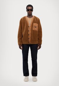 Cardigan en shearling marron avec une poche orange contrastante. Présente des boutons noirs, une coupe décontractée et un col rond. Porté avec un jean foncé.