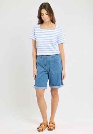 Femme portant une chemise à rayures bleues claires et blanches avec un col carré, un short en jean retroussé et des sandales marron avec boucle, debout devant un fond uni.