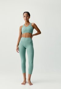 Svetlozelená športová podprsenka a legíny s vysokým pásom vyrobené z hladkého, elastického materiálu; podprsenka má krížový dizajn vpredu, oboje do dĺžky kolien.