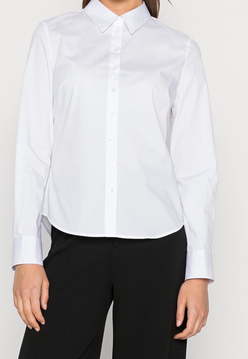 Hvid langærmet skjorte med knaplukning og klassisk krave, glat tekstur og afrundet kant, parret med sorte skræddersyede bukser.