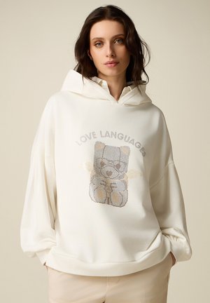 Jonge vrouw met een witte hoodie met een teddybeer en de tekst "LOVE LANGUAGES", staand met handen in de zakken tegen een effen achtergrond.