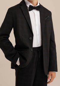 Černý kostkovaný blazer se dvěma knoflíky, nošený přes bílou košili. Zahrnuje černý motýlek a elegantní kalhoty s šňůrkou v pase.