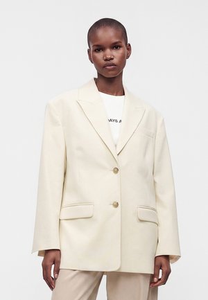 Νεαρό άτομο με κοντά μαλλιά φοράει oversized κρεμ σακάκι και ανοιχτόχρωμο παντελόνι πάνω σε απλό λευκό φόντο.