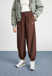 Hnědé volné kalhoty s elastickým pasem a ohrnutými kotníky. V kombinaci se světle zelenou bundou a bílými teniskami Nike s akcenty.