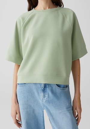 Felpa verde chiaro oversize con maniche raglan, collo a giro e orlo a costine. Texture liscia, abbinata a denim azzurro.