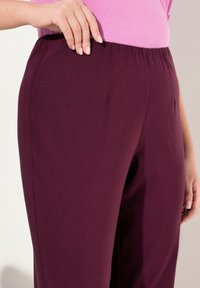 Bordeauxkleurige broek met een elastische tailleband, gemaakt van een gladde, rekbare stof en voorzien van zijzakken, gedeeltelijk zichtbaar met een hand rustend op de tailleband.