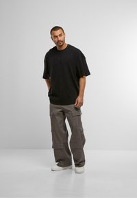 T-shirt noir oversize avec une finition texturée, associé à un pantalon cargo gris avec de nombreuses poches et une coupe décontractée. Baskets blanches.