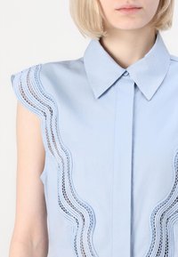 Camicetta senza maniche di colore azzurro chiaro con colletto a punta, caratterizzata da un bordo in pizzo scallop lungo la spalla. Tessuto liscio con parte anteriore a bottoni.