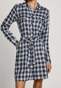 Abito a quadri navy e bianco con maniche lunghe, chiusura con bottoni, due tasche sul petto e una cintura legata in vita. Materiale in cotone con una superficie liscia.