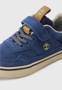 Sneaker en suède bleu avec une finition texturée, dotée d'une sangle Velcro, de lacets à motifs, et d'une semelle en caoutchouc beige avec un logo d'arbre sur le côté.