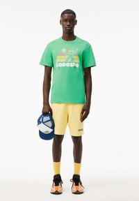 Camiseta de algodón verde con logo blanco, pantalones cortos amarillos, gorra azul y blanca, y zapatillas naranjas y negras. Diseño simple y deportivo.