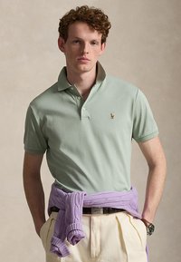 Νέος άνδρας με σγουρά μαλλιά που φοράει ανοιχτοπράσινο πόλο και μπεζ παντελόνια, με ένα μωβ πουλόβερ δεμένο γύρω από τη μέση.