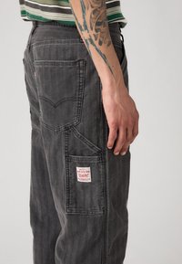 Pantalones de mezclilla gris oscuro con corte recto, que cuentan con múltiples bolsillos, detalles de etiqueta roja y una textura de rayas horizontales en la tela.
