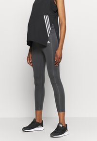 Černý sportovní top bez rukávů s bočními rozparky, v kombinaci s přiléhavými tmavě šedými legínami s kontrastním švem a logem Adidas. Černé tenisky.