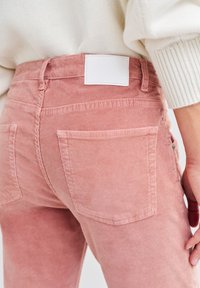 Roze corduroybroeken hebben een hoge taille, twee achterzakken en een wit label op de tailleband. De stof heeft een gestructureerde ribbelafwerking.