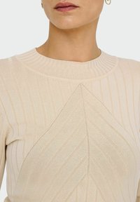 Kvinna som bär en beige ribbad tröja med ett chevron-mönster och gyllene hoopörhängen, visad från halsen till mitten av torse.
