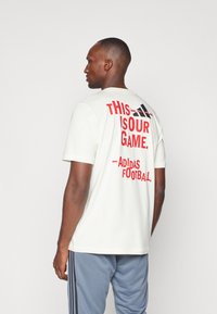 Hvid kortærmet T-shirt med rød tekst "DETTE ER VORES SPIL. - Adidas Fodbold." på ryggen, med rund hals og en afslappet pasform.